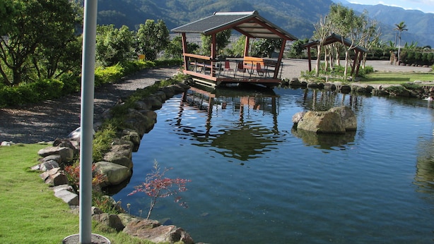 Un entorno tranquilo junto al lago con un pabellón de madera y una pequeña pérgola, rodeado de abundante vegetación y montañas al fondo.