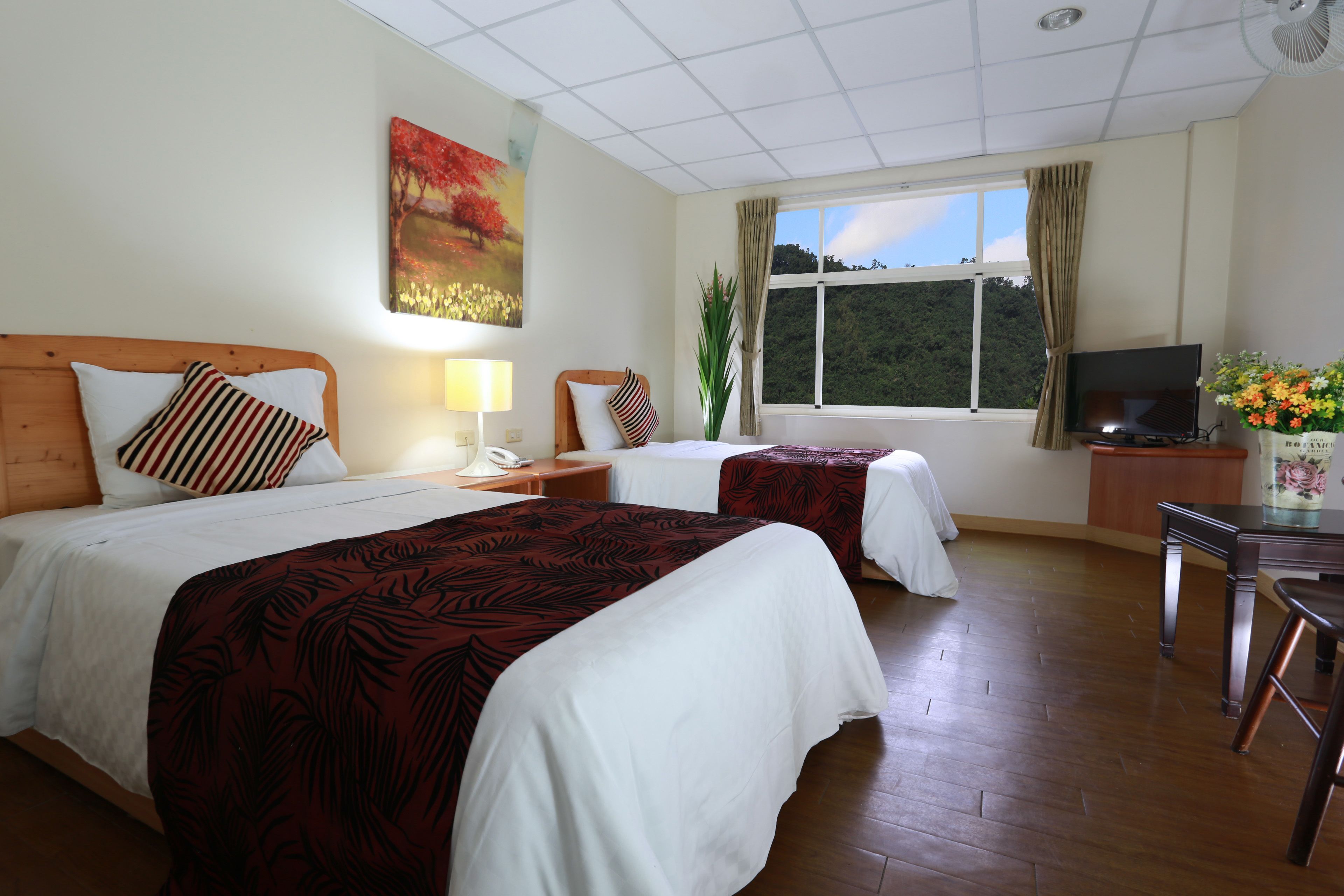 Habitación de hotel con dos camas, televisión, escritorio y ventana con vista a árboles.