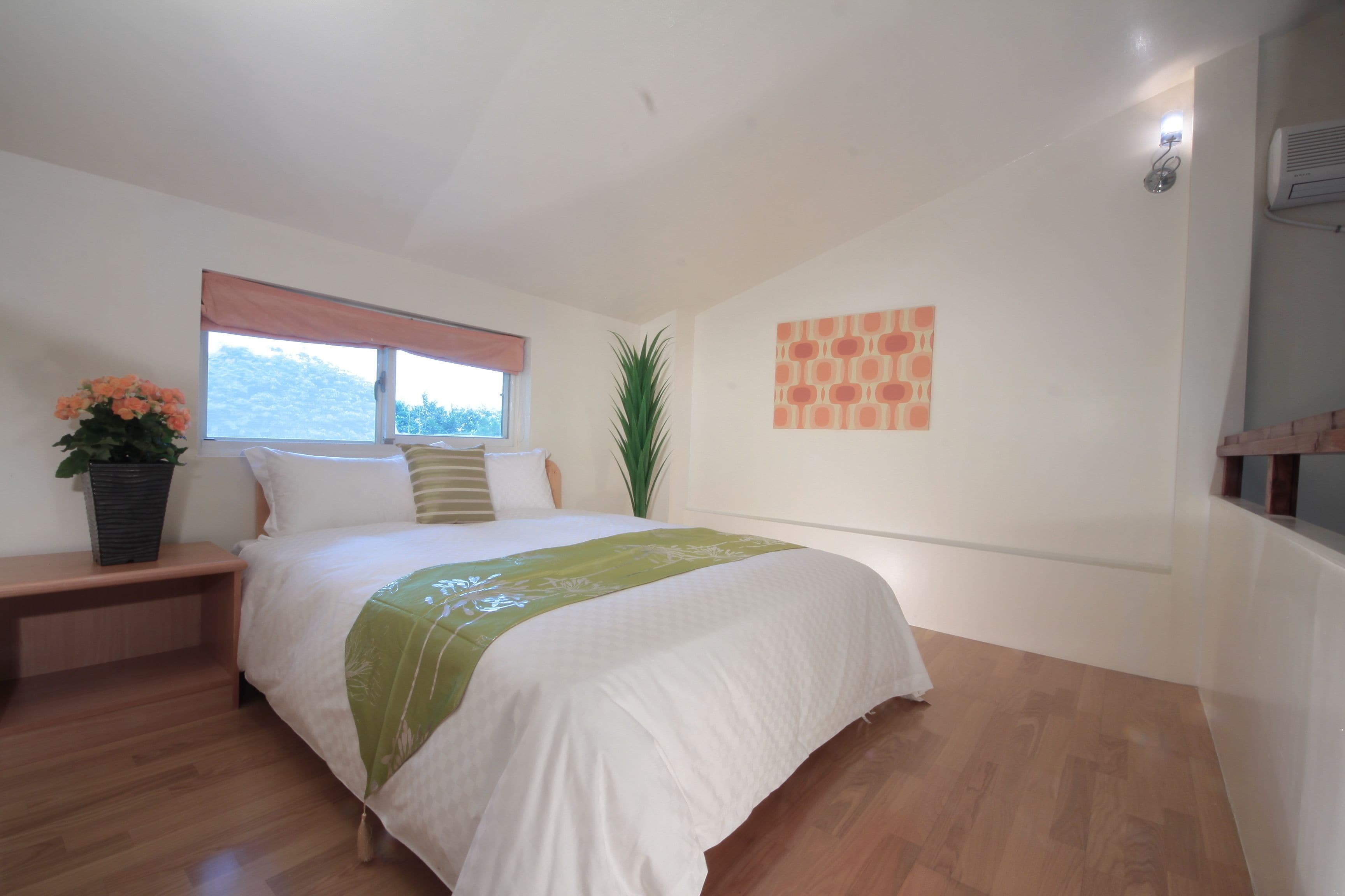 A bedroom with a bed, a nightstand, a window, a potted plant, and a wall-mounted air conditioner.