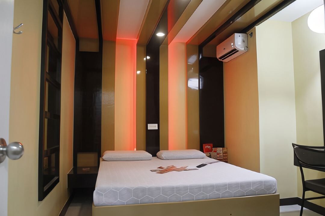 A narrow hotel room with a bed, a chair, and an air conditioning unit.