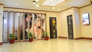 Un vestíbulo de hotel con suelo de baldosas, un mostrador de recepción, una pantalla de televisión y plantas en macetas.