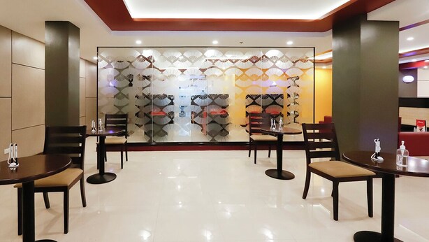 A modern restaurant with a central glass partition featuring a geometric pattern. Tables are arranged around the partition, each with chairs. The floor is tiled, and the ceiling has recessed lighting. - Hotel Sogo LRT Monumento Station (Caloocan)