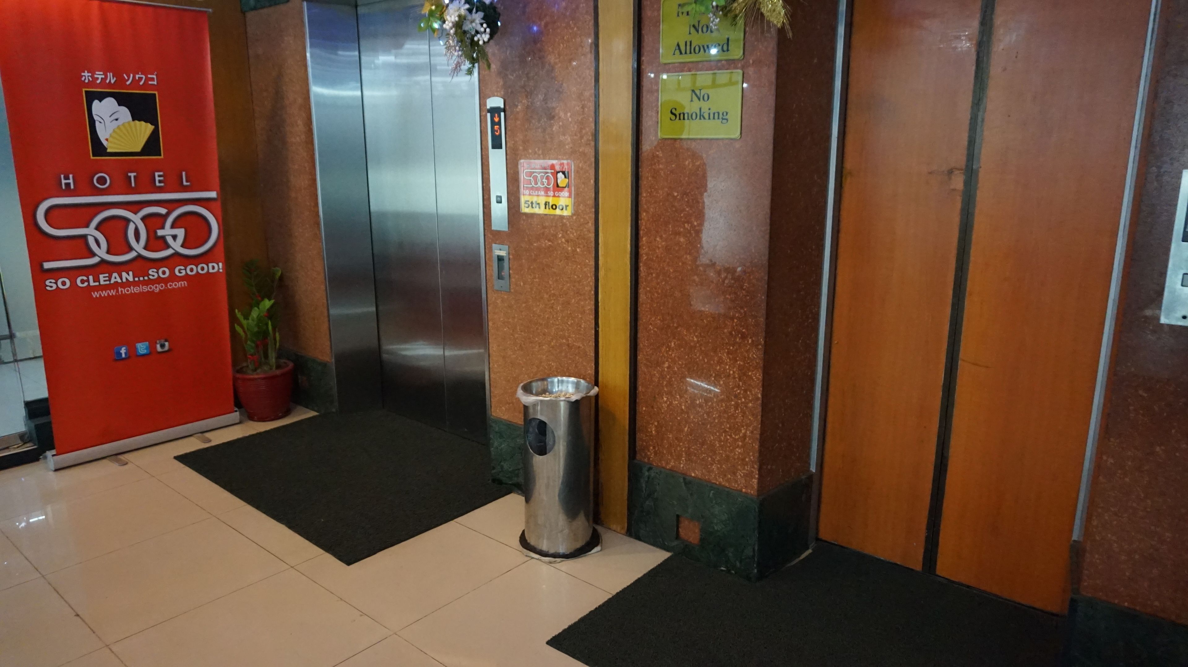 A hotel entrance with a red sign, an elevator, a trash bin, and a no-smoking sign.