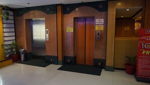 Zona de ascensores con dos elevadores, un cartel que indica prohibido fumar y un anuncio promocional para pedir comida y bebida dentro de un radio de 10 metros.