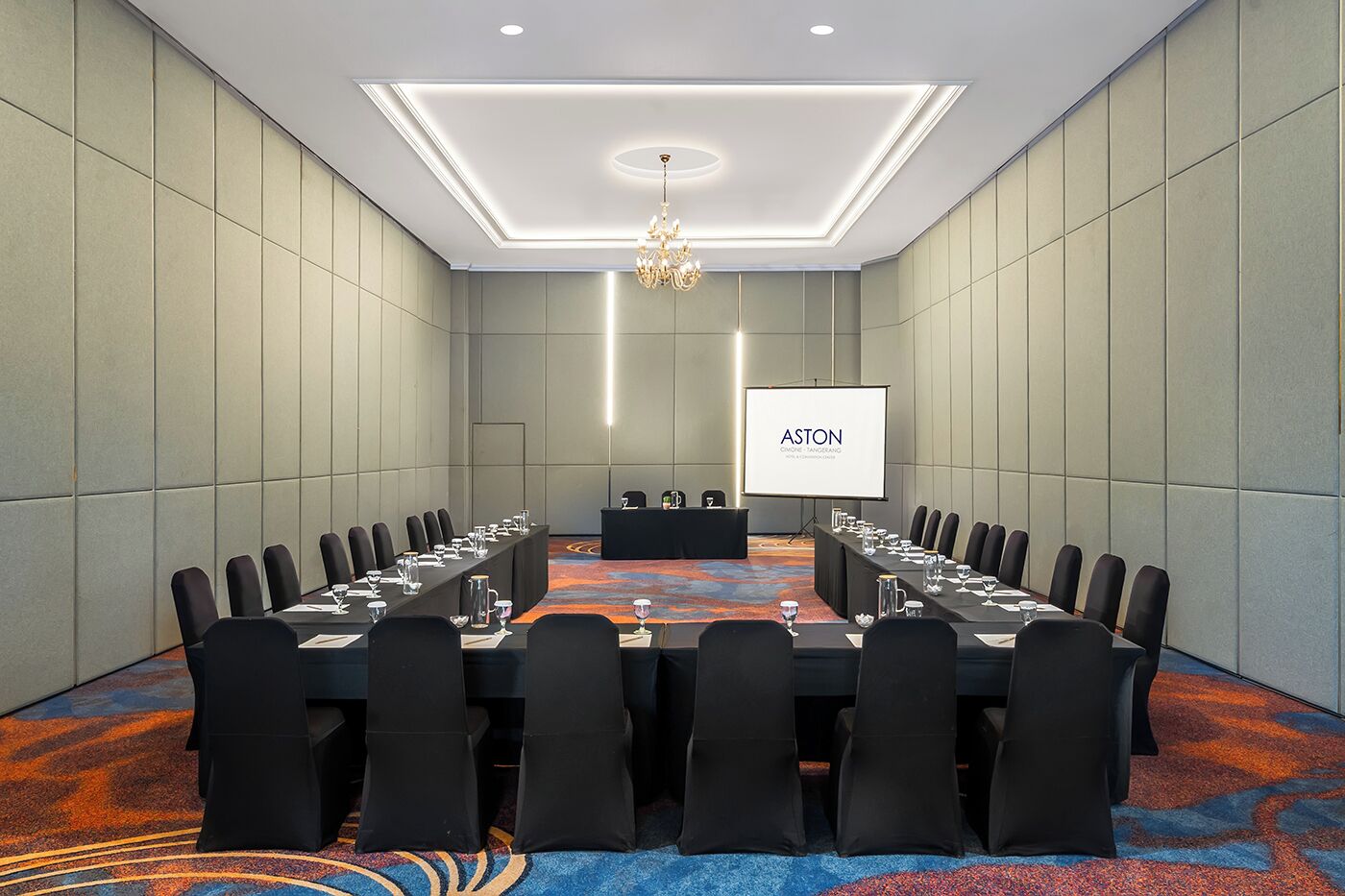 A conference room with a long table, black chairs, a projector screen, and a chandelier.