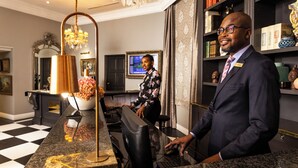 A man in a suit standing in a lobby with a desk, a computer, and a television. - The Fairlawns Boutique Hotel & Spa (Johannesburg)