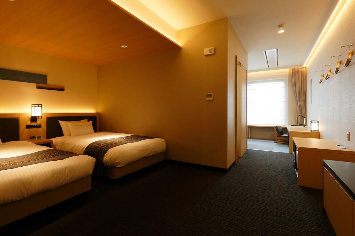 A hotel room with two beds, a desk, a chair, a window with curtains, and a wall-mounted air conditioning unit.
