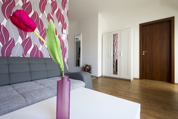 A modern living room with a grey sofa, a white coffee table, and a pink vase with a single flower. There is a wooden door and a mirror on the wall.