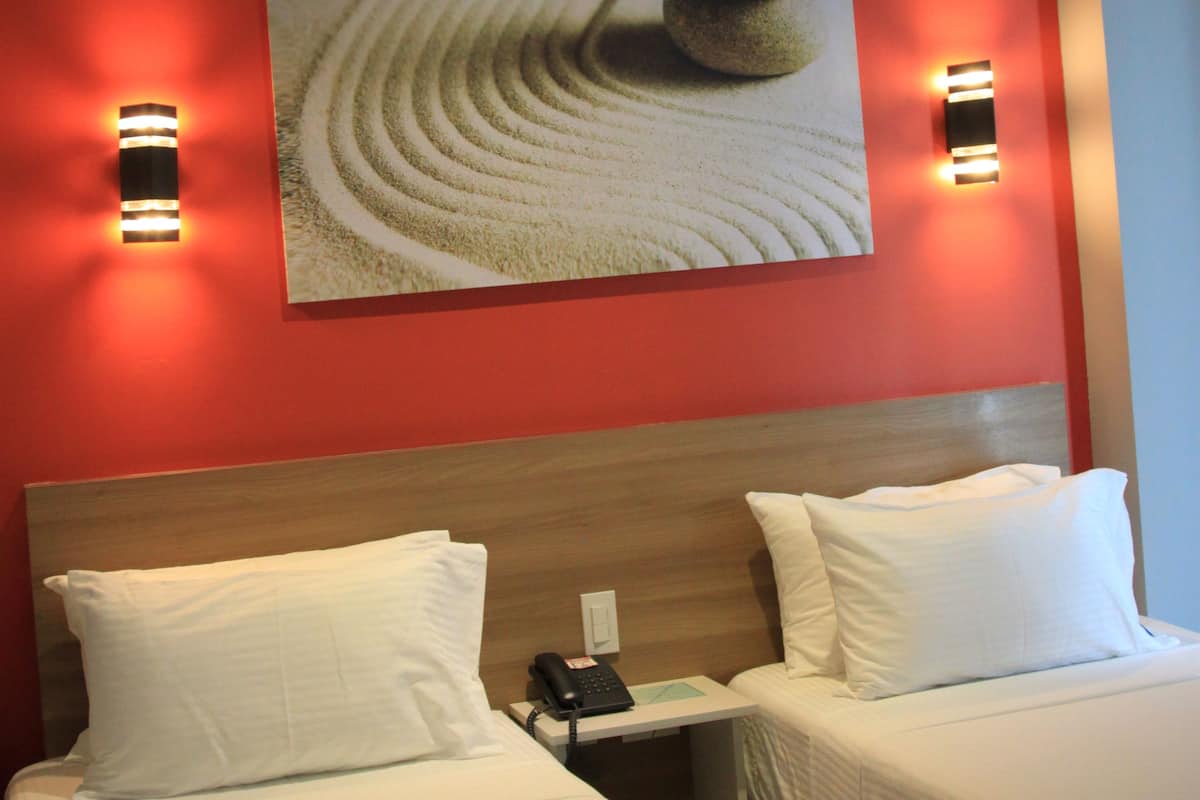 A hotel room with two beds, a wooden headboard, a painting on the wall, and wall-mounted lights.