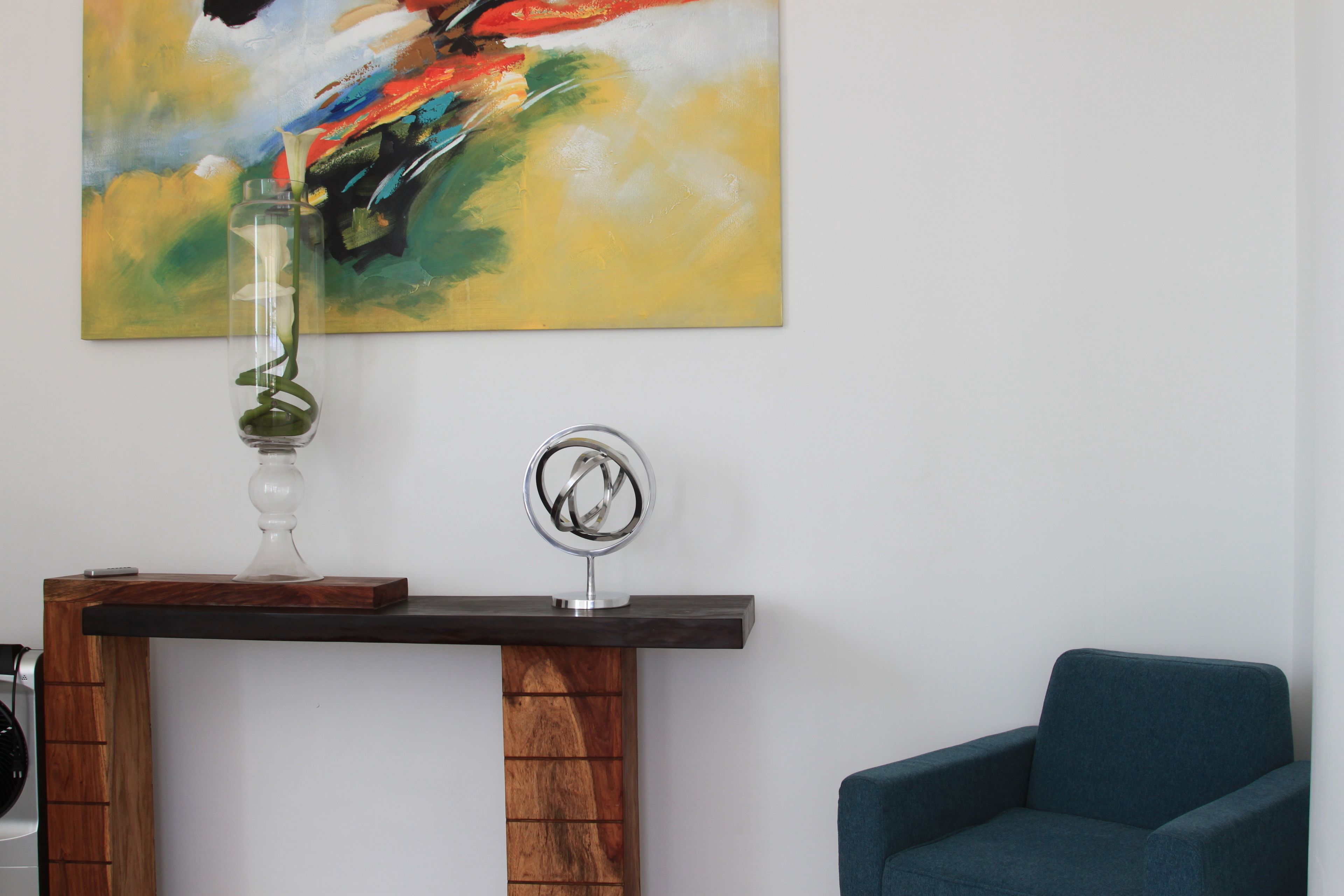 Una sala de estar moderna con una mesa consola de madera, un florero con una planta verde, un sillón azul y un cuadro abstracto colorido en la pared.
