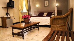 A bedroom with a bed, a wicker chair, a wooden table, a TV on a shelf, and a bedside lamp. - The Lusaka Hotel (Lusaka)