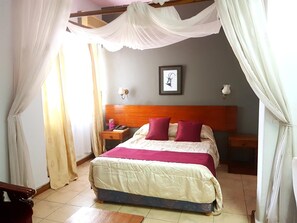 A bedroom with a bed, a nightstand, a wooden headboard, a framed picture on the wall, and sheer curtains. - The Lusaka Hotel (Lusaka)
