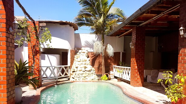 A swimming pool with a stone wall and palm trees, surrounded by a patio area with outdoor seating. - The Lusaka Hotel (Lusaka)