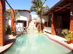 Une piscine bordée d’un mur en pierre et entourée de palmiers, avec un espace patio aménagé avec des sièges d’extérieur.