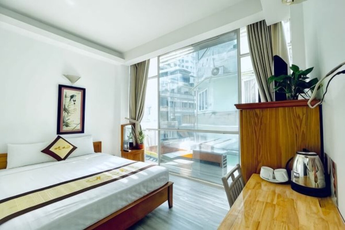 A hotel room with a bed, a wooden desk, a coffee maker, a plant, and a view of the city.