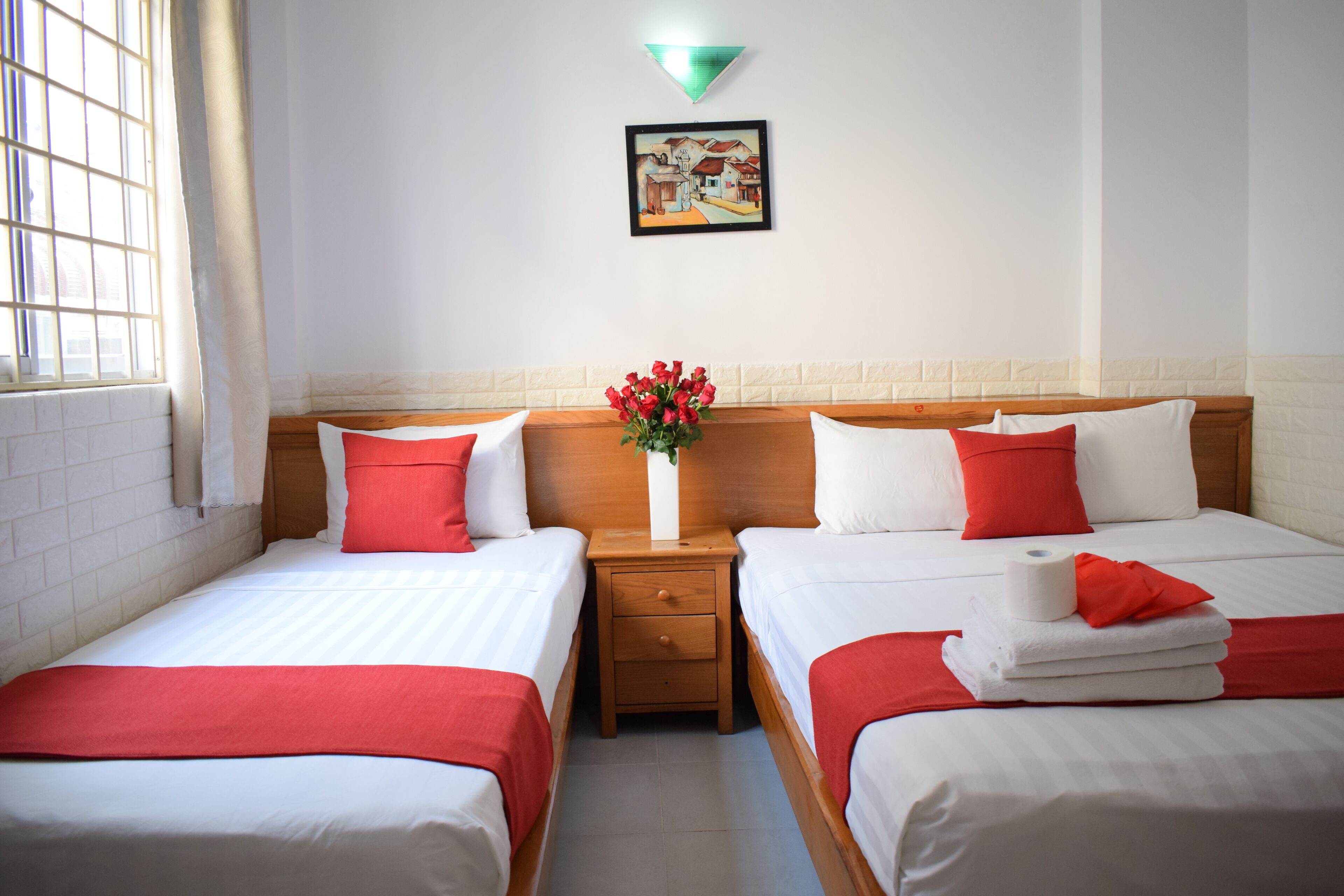 A hotel room with two beds, a nightstand, a vase with flowers, and a painting on the wall.
