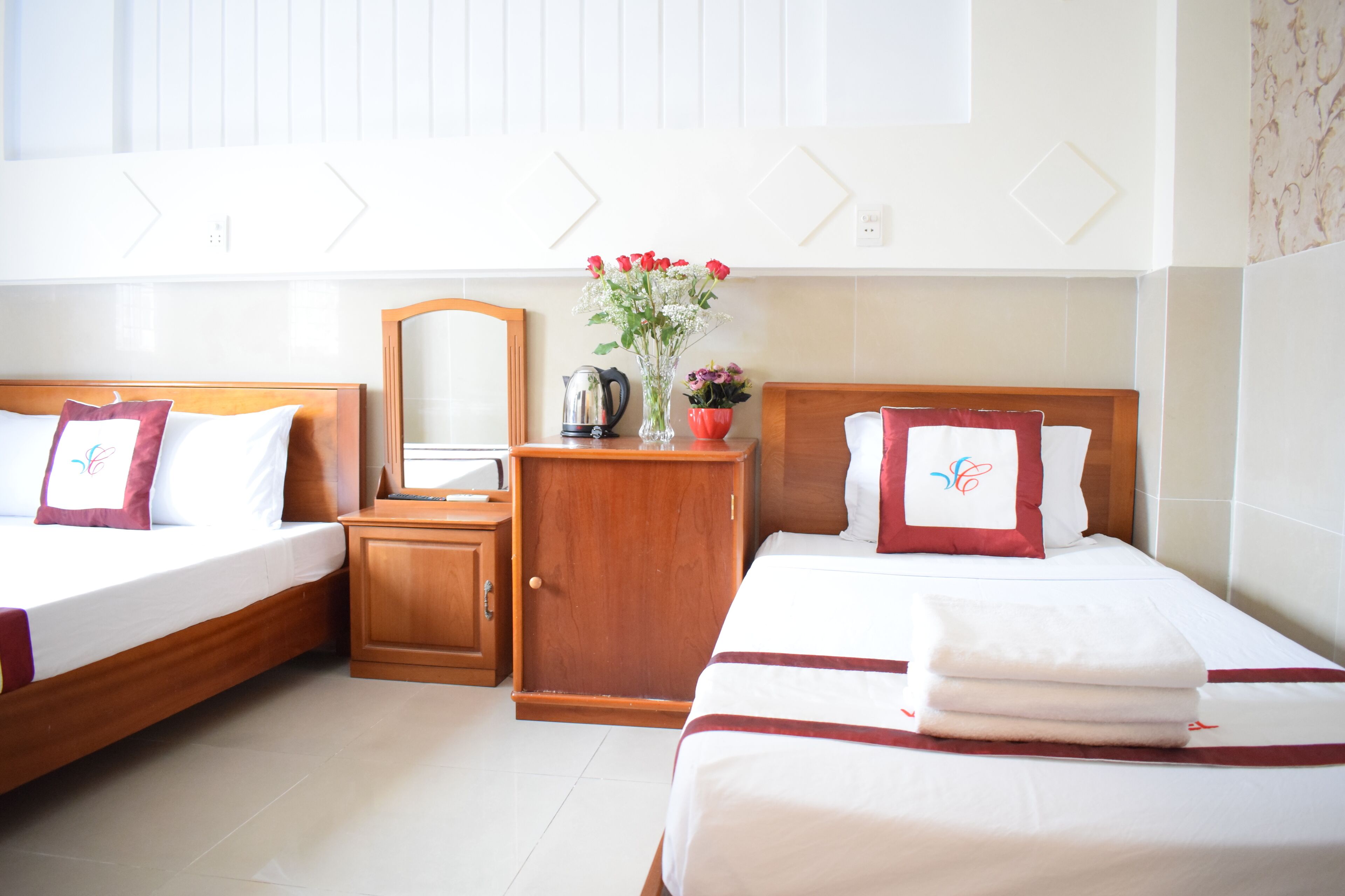 A hotel room with two beds, a wooden wardrobe, a mirror, a vase with flowers, and a kettle.