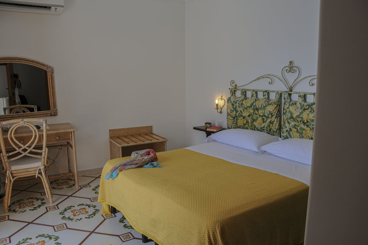 A room with a bed, a wooden desk, a chair, a mirror, and a nightstand with a lamp.