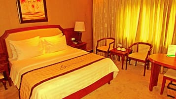 A hotel room with a large bed, two chairs, a small table, a nightstand, a lamp, and a painting on the wall.