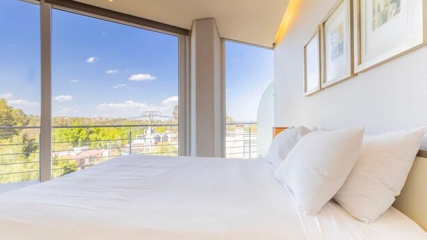 A bedroom with a large bed, white bedding, and a view of a cityscape through large windows.
