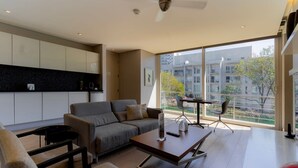 A modern living room with a grey sofa, a coffee table, and a dining area with chairs. There is a large window providing a view of buildings and trees.