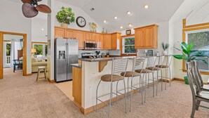 A modern kitchen with wooden cabinets, a stainless steel refrigerator, a microwave, and a bar area with stools. - Inn-Seaclusion: Elevator, Private Pool & Hot Tub, Theater Room (Corolla)