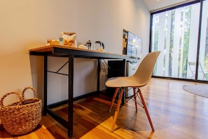 A modern office space with a wooden desk, a white chair, a wicker basket, and a large window with a view of trees.