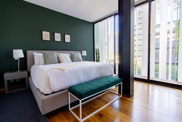 A modern bedroom with a large bed, a bench, and a view of buildings through large windows.