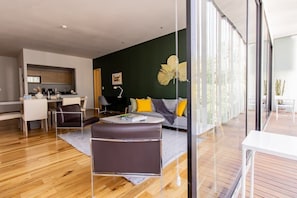 A modern living room with a sofa, coffee table, and chairs. There is a kitchen area with cabinets and a dining table. The room has large windows and a view of the outdoors.