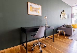 A modern living room with a dark grey wall, a black desk, a grey office chair, a small round table, and a grey sofa with a yellow pillow.