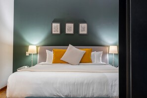 Een net opgemaakt bed met witte en gele kussens, een donkergroene muur en daarboven drie ingelijste foto's.