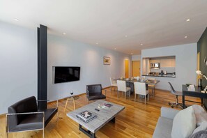 Una sala de estar moderna con un sofá, una mesa de centro, un televisor y un comedor con sillas, y una cocina al fondo.