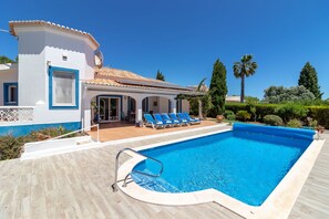 Une piscine avec une terrasse en bois, un bâtiment blanc orné de motifs bleus et un ciel d’un bleu limpide.