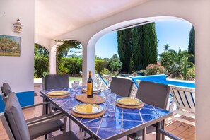 Un espace de restauration en plein air avec une table dressée pour deux personnes, une bouteille de vin et une vue sur une piscine et un jardin.