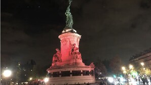 A nighttime view of a large, illuminated monument with a statue on top, surrounded by a plaza with people and streetlights.
