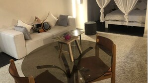 A modern hotel room with a glass coffee table, a sofa with cushions, a bed with white bedding, and a canopy bed.
