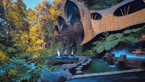 A unique treehouse structure with a wooden exterior and circular openings, surrounded by lush greenery and a small pond with stepping stones. - Sanna Ubud A Pramana Experience (Tegallalang)