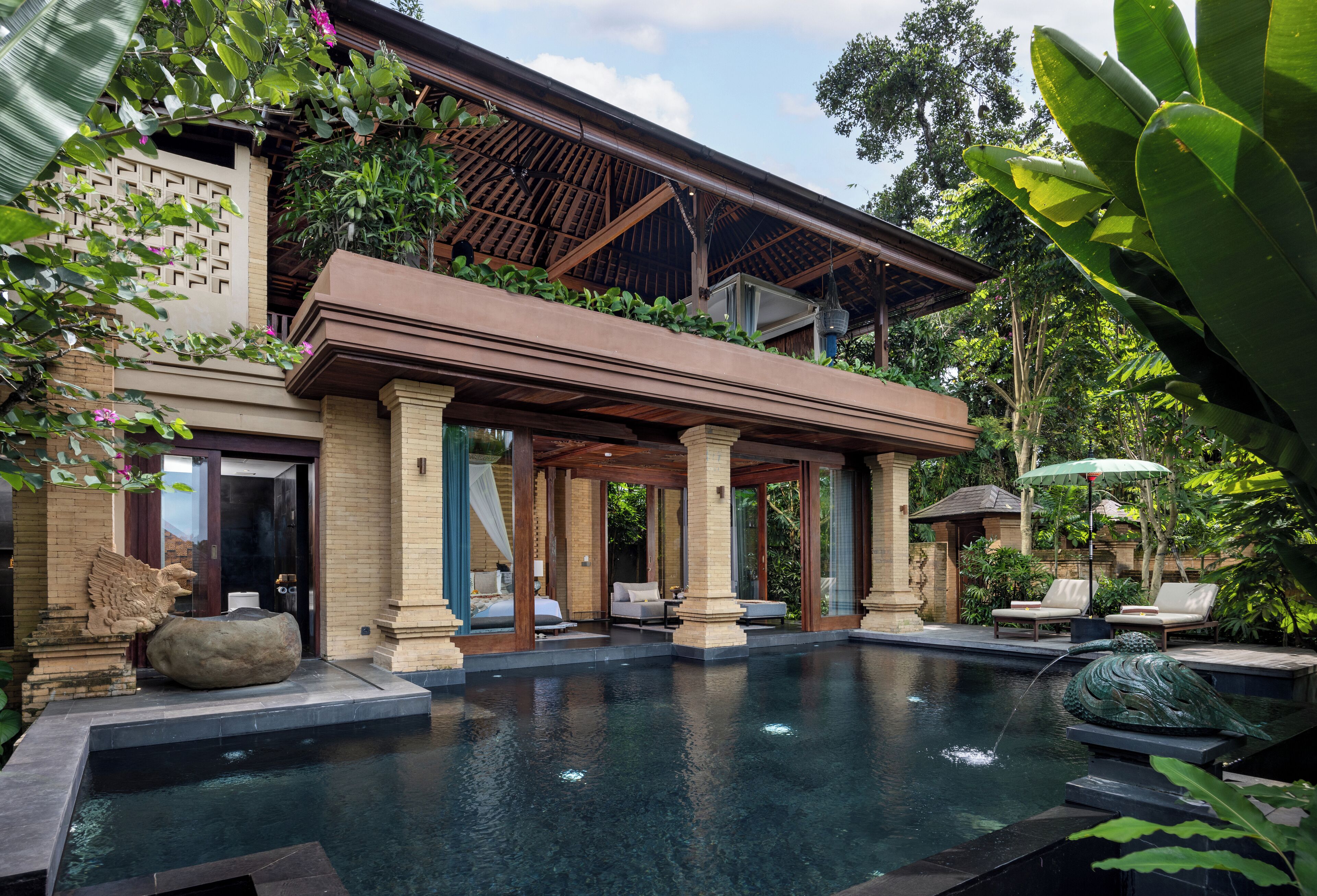 A luxurious pool area with a covered seating area, stone sculptures, and lush greenery.