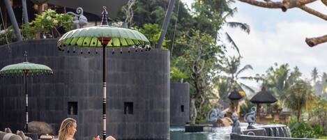 A poolside area with lounge chairs and umbrellas, a stone wall, and a water feature with statues.