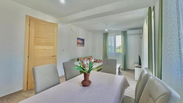 A modern room with a dining table, chairs, a wooden door, a painting on the wall, and a view of another room through a window. - Talia Apartments (Lastva Grbaljska)