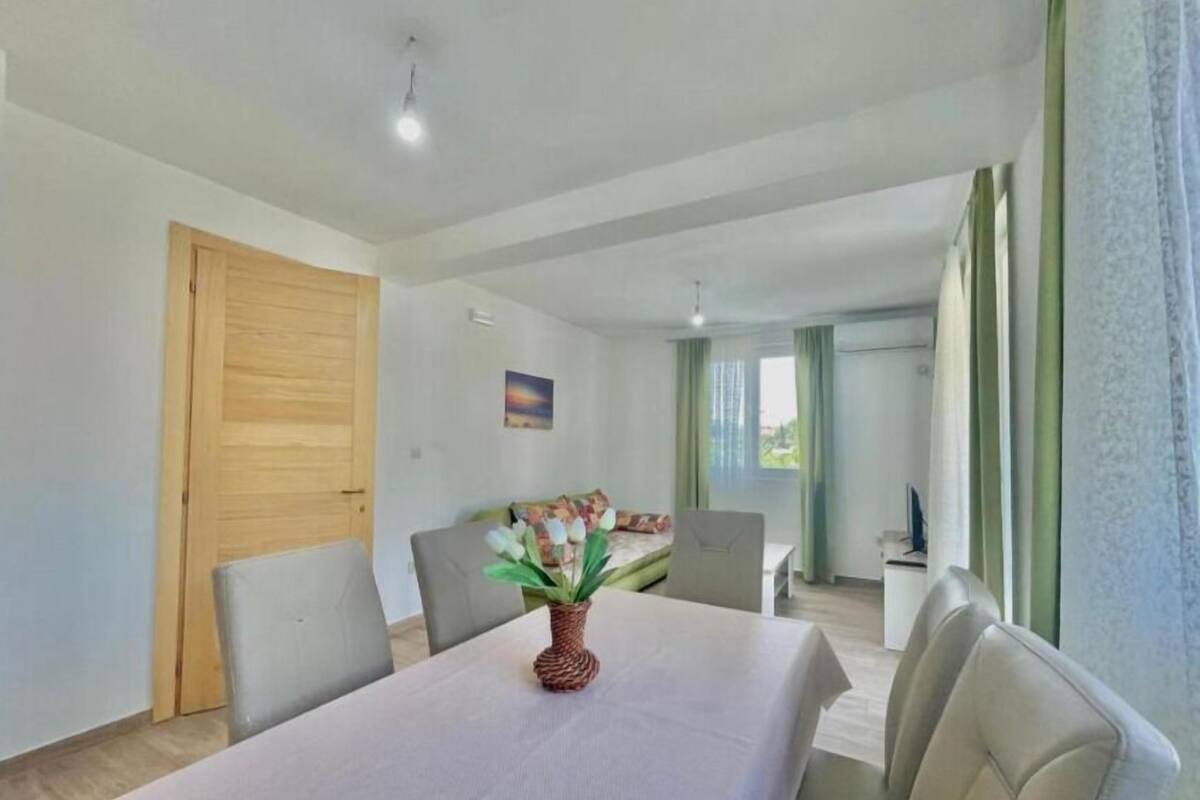 Ein modernes Zimmer mit Esstisch, Stühlen, einer Holztür, einem Bild an der Wand und Blick in einen anderen Raum durch ein Fenster.