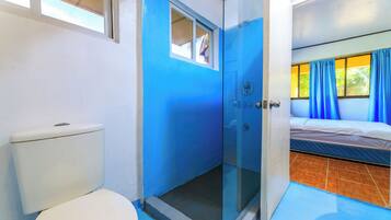 A bathroom with a toilet, a glass-enclosed shower, and a bed with blue curtains.