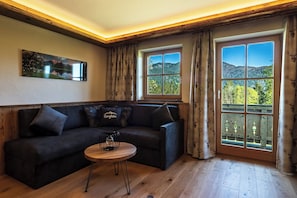 Ein gemütliches Zimmer mit einem dunkelgrauen Sofa, einem kleinen Holztisch und einem großen Fenster mit Blick auf eine bewaldete Landschaft.
