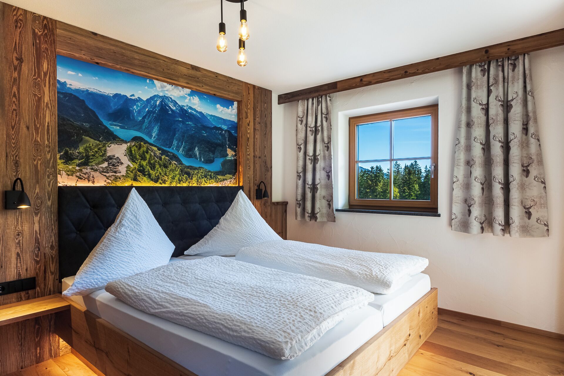 Een slaapkamer met een groot bed, een houten hoofdbord, een schilderij van bergen, een raam met gordijnen, en een nachtkastje met een lamp.