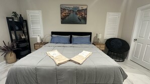 A neatly made bed with a grey bedspread and two folded towels, a black chair, a bedside table with a lamp, a framed picture on the wall, and a potted plant. - Guesthouse near Anaheim Convention (Santa ana)