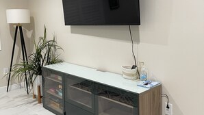 A modern living room with a flat-screen TV mounted on the wall, a glass-fronted entertainment unit, a floor lamp, and a pair of slippers on the floor. - Guesthouse near Anaheim Convention (Santa ana)
