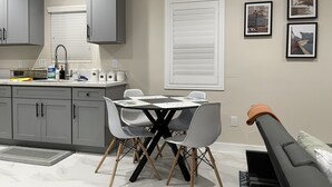 A modern kitchen with grey cabinets, a dining table with chairs, and a window with blinds. - Guesthouse near Anaheim Convention (Santa ana)