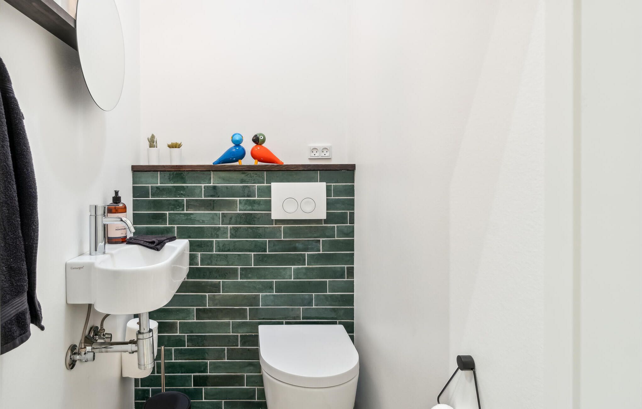 A bathroom with a white sink, a toilet, and a green-tiled wall. There are two colorful bird figurines on the wall, a hand soap dispenser, and a towel hanging on the wall.
