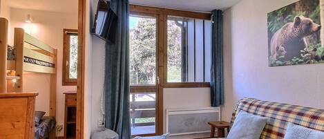 A cozy room with a wooden floor, a plaid sofa, a small table, a TV mounted on the wall, and a window with curtains.