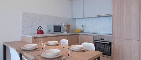 A modern kitchen with a wooden dining table set for four, white chairs, a microwave, a kettle, and a stove.